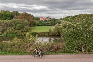Zwei Frauen fahren auf dem alsphaltierten Saaleradweg mit dem Fahrrad. Neben ihnen fließt der Fluss Saale. Im Hintergrund sieht man Schloss Goseck auf den Hügel.