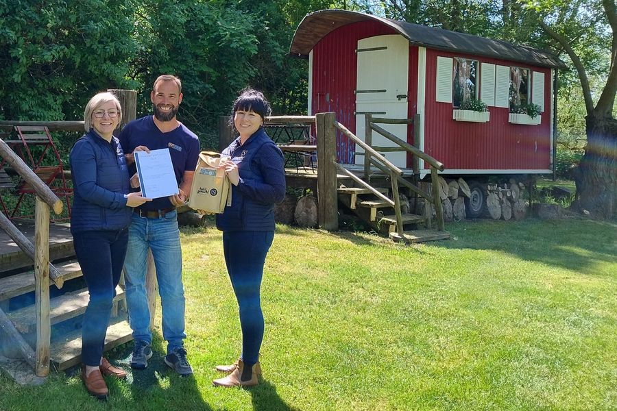 Zwei Frau und ein Mann stehenauf einer grünen Wiese, im Hintergrund ist ein Schöferwagen zu sehen. Sie halten eine Urkunde und eine braune Tasche hoch (v.l. Antje Peiser, Geschäftsführerin Saale-Unstrut Tourismus GmbH / Jens Bellmann (Inhaber OUTTOUR) / Carolyn Fox, Projektkoordinatorin Produktcheck Saale-Unstrut Tourismus GmbH)