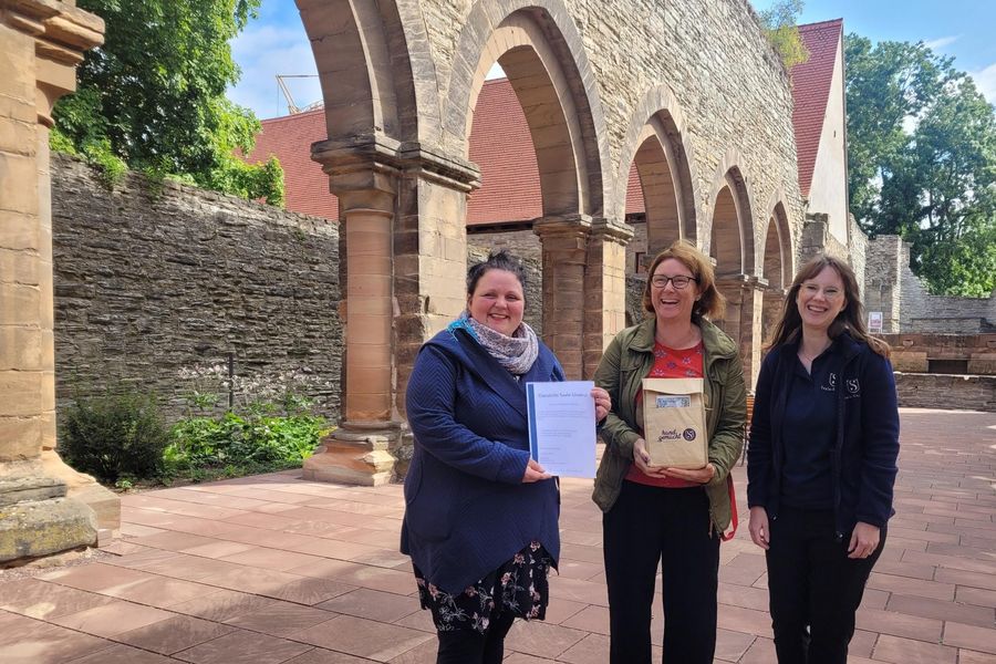 Drei Frauen stehen lachend nebeneinander. Die linke Frau hält eine Urkunde in der Hand, die mittlere eine kleine Tasche mit regionalen Produkten. Dahinter sind Spitzbögen einer Klosterruine zu sehen. Die Sonne scheint.